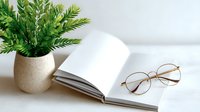 Open blank notebook with round gold-framed reading glasses and decorative green plant in ceramic pot on white surface, minimalist workspace setup.