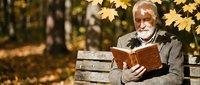 Senior Caucasian man with white beard reading book on park bench surrounded by golden autumn maple leaves creating peaceful outdoor learning moment.