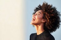 Young African American woman with natural curly hair laughing joyfully against soft sunlit background, expressing genuine happiness and positive emotions.