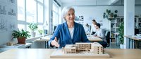 Senior female architect with gray hair and South Asian ethnicity presenting wooden architectural model at desk in modern office with colleagues working in background.