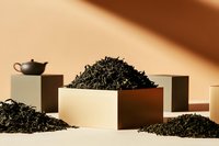 Loose black tea leaves arranged on minimal geometric platforms with traditional teapot, creating modern composition against warm beige background.