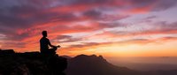 Silhouetted figure meditating on mountain peak at dramatic sunset with vibrant orange and purple sky, birds flying overhead, and layered mountain ranges below. Wellness and mindfulness concept.