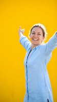Cheerful middle-aged woman with blonde hair wearing light blue shirt with arms raised in celebration against bright yellow background, expressing joy and success.