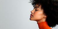 Young African American woman with natural afro hair in orange turtleneck sweater, eyes closed in serene profile pose against light background. Minimalist beauty portrait.