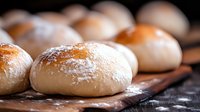 Fresh raw bread dough balls dusted with flour on wooden board, close up view with selective focus showing soft texture and artisanal baking preparation.