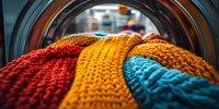 Colorful knitted wool sweaters in red, yellow and turquoise seen through round washing machine door glass. Close up view of laundry in washer drum.
