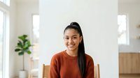 Young woman with long dark hair and warm smile wearing rust-colored ribbed sweater, seated indoors with bright natural light and modern home decor in background. Suitable for lifestyle, wellness, and portrait projects.