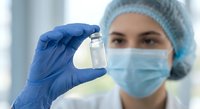 Female healthcare worker in protective mask and cap holding medical vial in laboratory. Professional medical research and pharmaceutical development concept.