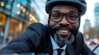 Cheerful African American businessman in glasses and bike helmet taking selfie on urban street with bokeh lights and buildings in background.