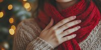 Woman wearing cozy knit sweater and red scarf against blurred Christmas lights background, creating warm holiday atmosphere and winter comfort feeling.