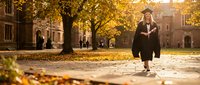 Young woman graduate in black cap and gown walking on university campus courtyard during golden autumn sunset with fallen leaves scattered on ground.