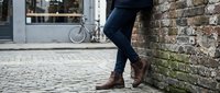 Stylish woman in slim jeans and brown leather ankle boots leaning against a rustic brick wall on a cobblestone urban street.