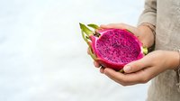 Close-up of hands holding fresh pink dragon fruit with green leaves against neutral background. Tropical fruit for healthy eating, nutrition, and culinary use.