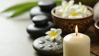 Spa wellness scene with burning candle, hot stones, plumeria flowers and coconut bowl for relaxation massage therapy and aromatherapy treatment sessions.
