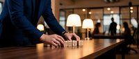 Businessman in navy suit arranging wooden blocks with house symbols on wooden desk in modern office with warm lighting, representing real estate planning and property investment strategy.