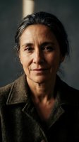 Middle-aged woman with dark hair and olive complexion wearing brown collared shirt, studio portrait with dramatic side lighting against neutral background.