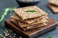 Crispy multigrain crackers with seeds stacked on wooden cutting board, fresh herbs and scattered pumpkin seeds on dark background, healthy snack concept.