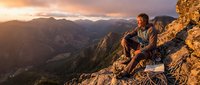 Male hiker sits on rocky mountain peak during golden hour sunset, overlooking dramatic valley landscape. Adventure travel and outdoor recreation concept.