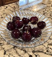 Fresh dark red cherries with stems arranged in crystal glass bowl on marble counter, highlighting natural gloss and texture of ripe fruit. Food photography setting.