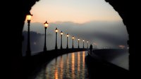 Romantic waterfront promenade with vintage lampposts at sunset, viewed through stone archway. Warm golden light reflects on calm water creating peaceful atmosphere.