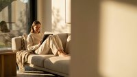Young woman relaxing on modern sofa in bright living room with natural sunlight streaming through large windows creating warm shadows on wall.