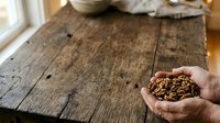Unrecognizable person holding a handful of granola mixture on rustic wooden table surface with natural window light for food styling and nutrition content.