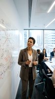 Professional mixed-race businesswoman presenting strategy on whiteboard during corporate meeting in modern office conference room with city view.