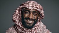 Middle Eastern man wearing traditional red and white keffiyeh headdress smiling warmly against dark gray background.
