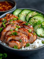 Grilled teriyaki chicken slices on steamed rice with fresh cucumber, green onions and sesame seeds in dark bowl. Spicy sauce in background on stone surface.