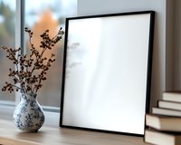 Empty black frame mockup on wooden table with oriental blue and white vase containing dried flowers, stack of books in background, natural window light.