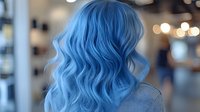 Young woman with vibrant blue wavy hair styled in long loose curls shown from behind in salon setting with soft bokeh lights background.