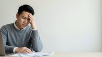 Asian male professional experiencing stress or fatigue while reviewing documents at desk, suitable for business, mental health, and workplace wellness concepts.
