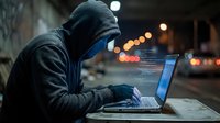 Hooded figure in dark clothing working on laptop computer at night on urban street with bokeh lights creating mysterious cybercrime atmosphere.