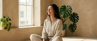 Young woman in beige loungewear meditating in sunlit room with houseplants. Peaceful morning wellness routine for mental health and mindfulness practice.