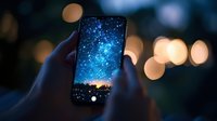 Smartphone screen displaying starry night sky and silhouetted treeline against vibrant blue galaxy background with blurred bokeh lights in foreground.