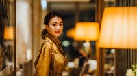 Young Asian businesswoman in elegant golden blazer smiling confidently in upscale hotel lobby with warm ambient lighting for corporate hospitality concepts.