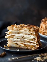 Layered crepe cake with caramel cream filling and crushed nuts on top served on dark plate against moody background, close up view of dessert slice.