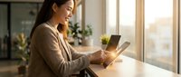 Young Asian businesswoman using digital tablet at modern office desk near window with natural sunlight for remote work and productivity.