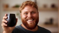 Young bearded redhead man with bright smile holding black coffee mug in cozy home setting. Warm atmospheric lighting creates welcoming morning mood.