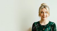 Young woman with blonde hair and fair complexion wearing dark green floral top, smiling at camera against neutral background. Suitable for business, lifestyle, and portrait projects.