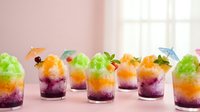 Colorful layered cocktails with vibrant green, orange, and purple gradients, garnished with fresh mint and decorative cocktail umbrellas in glasses against a soft pink background, perfect for beverage, bar, and hospitality design projects.