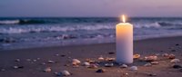 White candle burning on sandy beach at sunset with seashells scattered around, ocean waves in background creating peaceful meditation atmosphere.