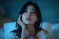 Young Asian woman with dreamy expression in moody blue lighting, touching face gently while resting on bed, portraying sensuality and contemplation.