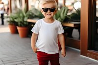 Cheerful young boy in white t-shirt and red pants wearing sunglasses, standing outdoors with confident smile against blurred cafe terrace background with potted plants.