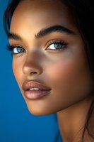 Close-up portrait of young mixed-race woman with glowing skin and blue eyes against vibrant blue background for beauty and skincare campaigns.