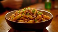 Crispy stir-fried potato strips garnished with sesame seeds, chili flakes and fresh green onions in dark ceramic bowl on wooden table. Asian fusion cuisine.