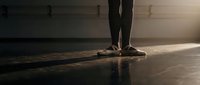 Ballerina in pointe shoes standing on wooden stage floor with dramatic warm lighting, casting long shadows during rehearsal or performance preparation.
