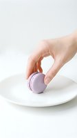Hand picking a lavender macaron from a white plate, minimalist food styling with soft light, pastel purple French cookie on clean background.