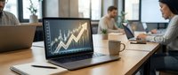 Laptop displaying financial growth chart with upward trending graph in modern office workspace with business professionals collaborating in background.