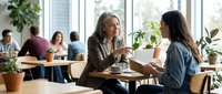 Two professional women having business meeting in modern cafe discussing project over coffee with colleagues working in background.